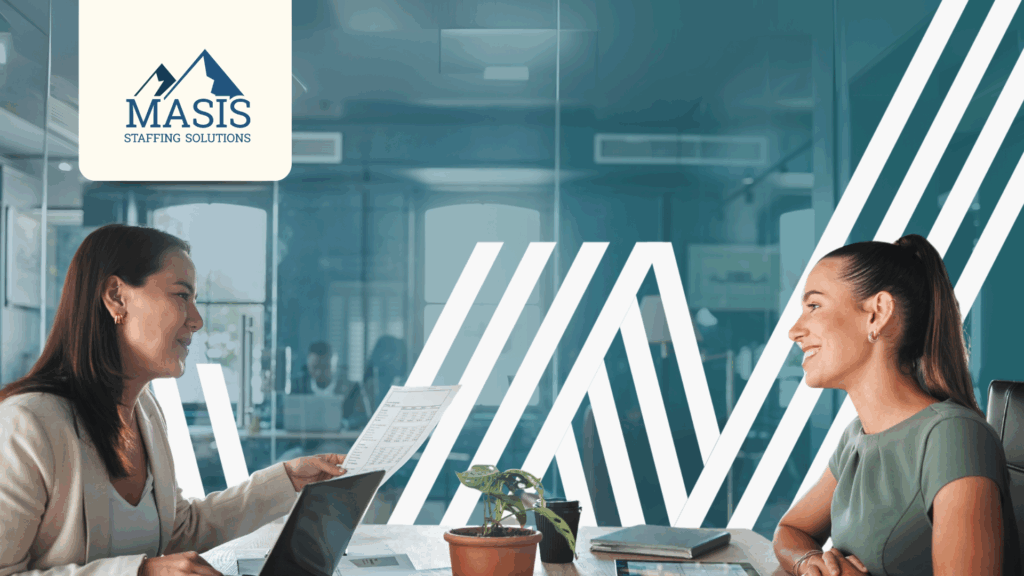 An interviewer and job seeker speak over a desk with various gadgets and potted plants