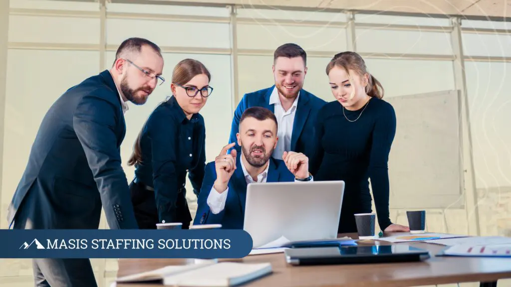 A group of engaged professionals and recruiters gathers around a laptop in collaborative discussion
