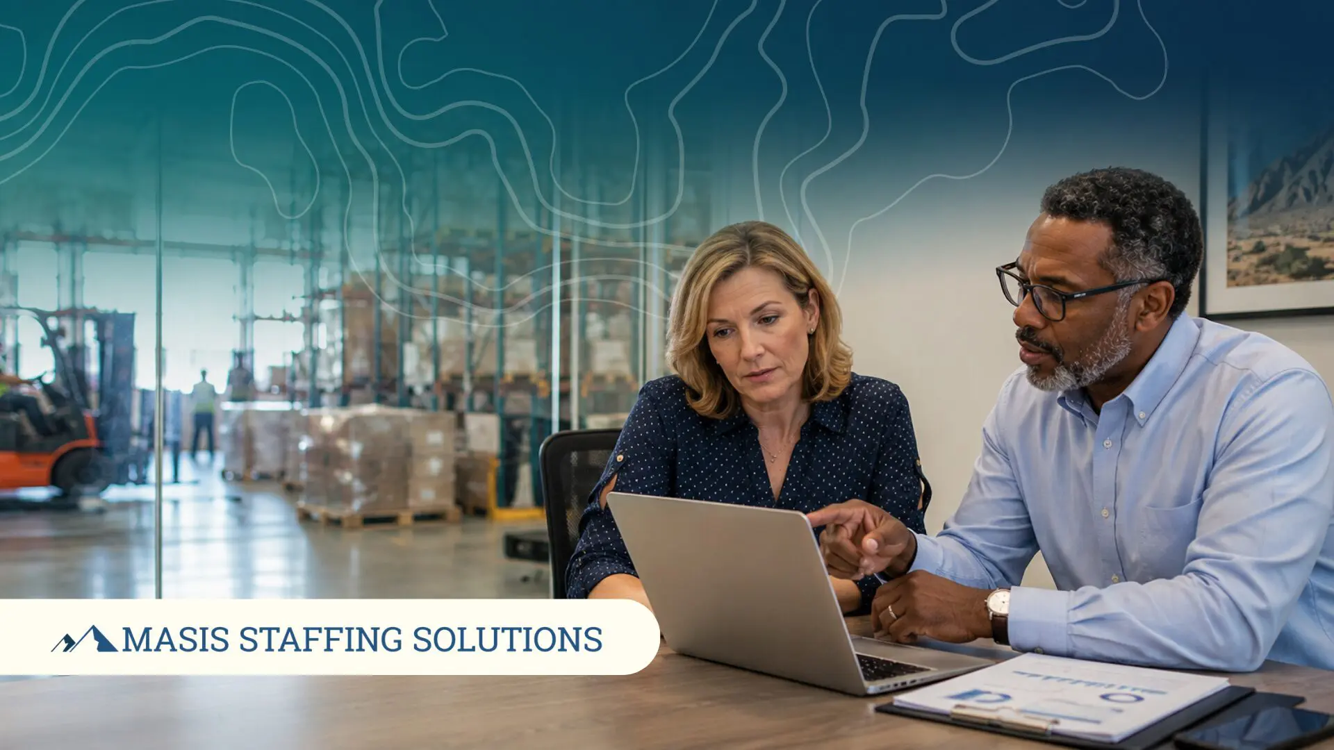 An employer and colleague evaluate staffing partner performance together on a laptop in a warehouse office