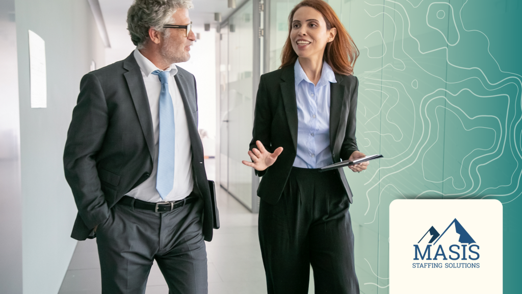 Woman in corporate attire walks down an office hallway with her manager, speaking enthusiastically